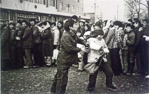 排队买彩票。彩票在80年代农村是一个新鲜词，只有在富裕的镇上才会有那么一家，图中为一位插队买彩票的农妇被彩票站工作人员推了出来。